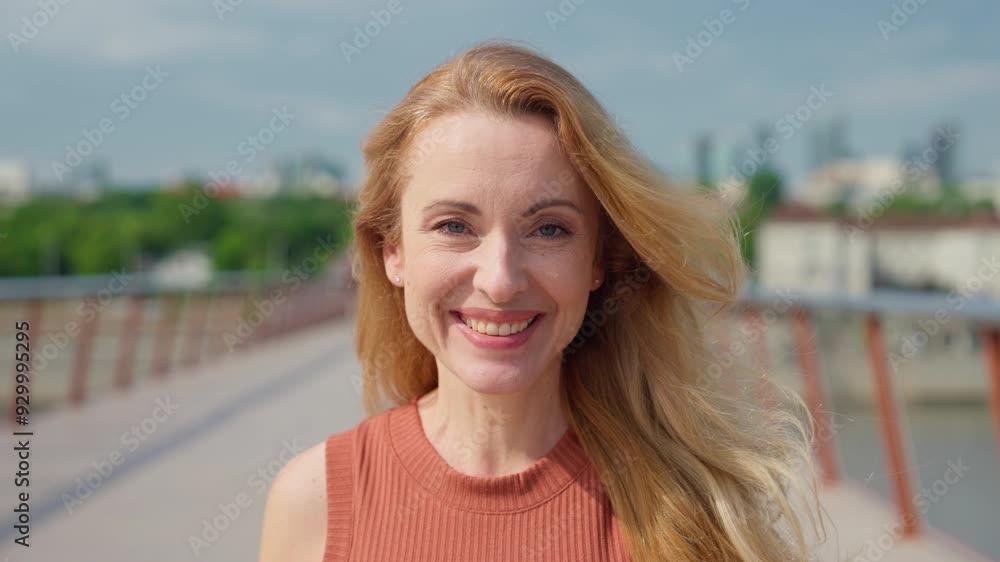 Cute Caucasian woman glancing directly at camera. Smiling widely while standing on long bridge. Female fixing her long blonde hair. Enjoying trip to new city or location. Positive attitude. Cute Caucasian woman glancing directly at camera. Smiling widely while standing on long bridge. Female fixing her long blonde hair. Enjoying trip to new city or location. Positive attitude.