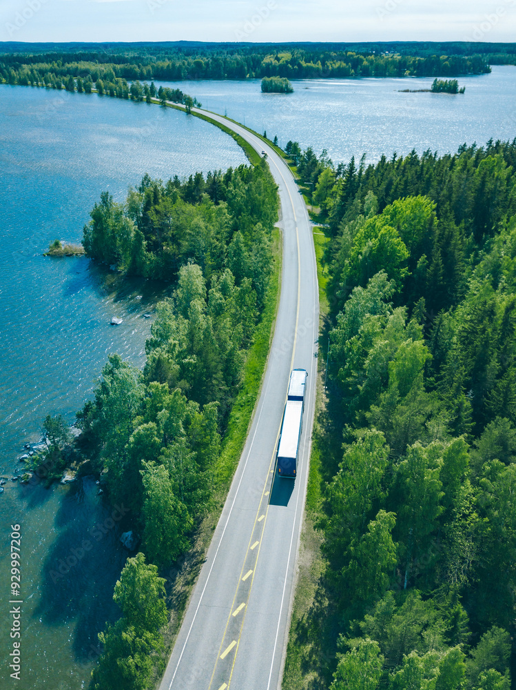 Aerial view of road with truck car in green woods by blue lakes sea water in Finland