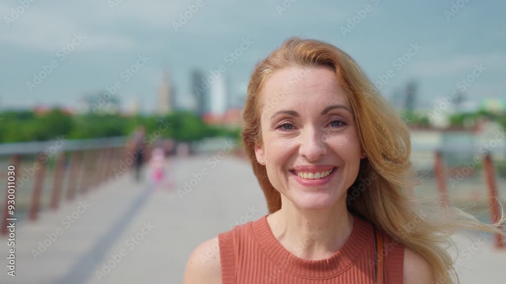 Pretty Caucasian woman glancing directly at camera and smiling widely. Positive emotions. Green trees visible in distant blurred background. Female relaxing during summer vacation in new location. Pretty Caucasian woman glancing directly at camera and smiling widely. Positive emotions. Green trees visible in distant blurred background. Female relaxing during summer vacation in new location.