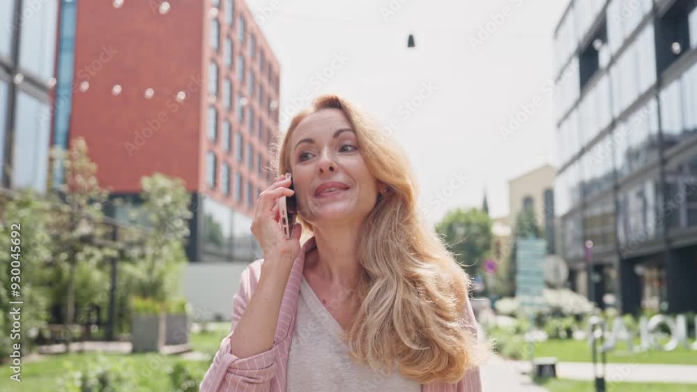 Happy Caucasian woman walking in middle beautiful street on sunny day. Holding smartphone while remotely talking with her friend or relative. Having positive mood while enjoying outside world. Happy Caucasian woman walking in middle beautiful street on sunny day. Holding smartphone while remotely talking with her friend or relative. Having positive mood while enjoying outside world.