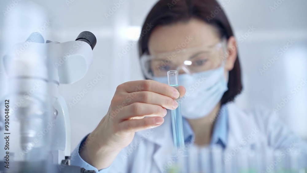 Scientist female researcher with protective glasses and mask examining lab tube with a blue fluid. Microbiology and science concept Scientist female researcher with protective glasses and mask examining lab tube with a blue fluid. Microbiology and science concept