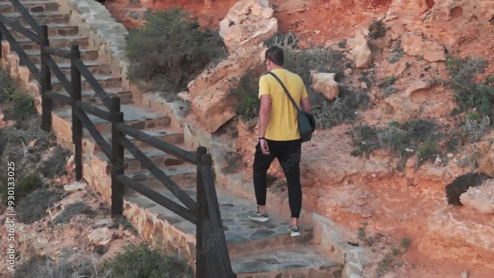A man in black pants and a yellow shirt descends stone steps by the ocean at sunrise, carrying a bag over his shoulder, recorded in slow motion. A man in black pants and a yellow shirt descends stone steps by the ocean at sunrise, carrying a bag over his shoulder, recorded in slow motion.