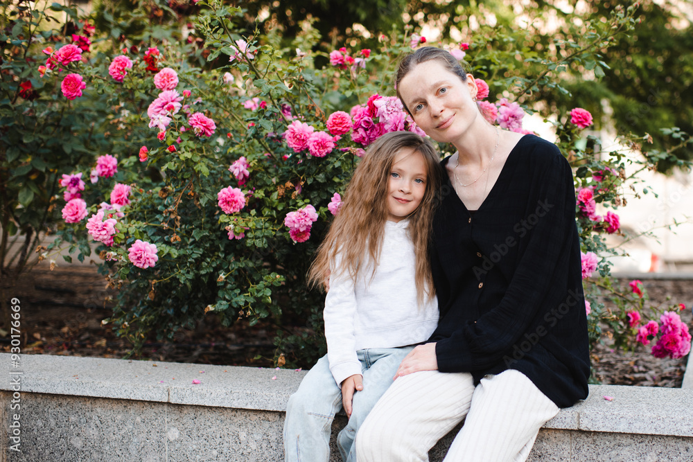 Fototapeta premium Young smiling woman holding child girl 5-6 year old over blooming rose flowers at city street outdoor. Childhood.