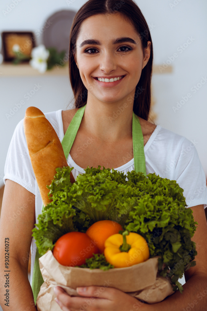 An attractive young dark-haired woman holding the paper bag full of vegetables while standing and smiling near the kitchen desk. Cooking and householding concepts An attractive young dark-haired woman holding the paper bag full of vegetables while standing and smiling near the kitchen desk. Cooking and householding concepts