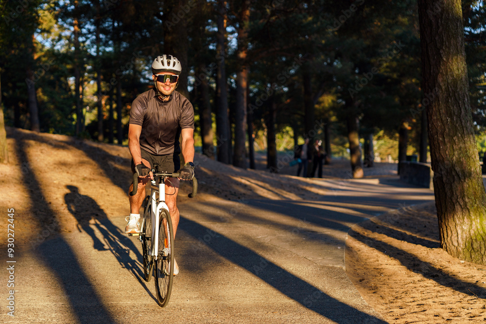 Fototapeta premium A cyclist in a helmet and sunglasses rides along a sun-dappled forest path, casting a long shadow.
