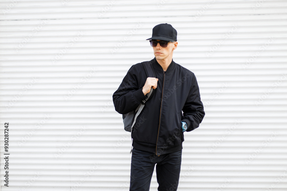 Cool handsome man with cap with sunglasses in fashion black outfit with jacket with backpack walks on the street near a white metal background Cool handsome man with cap with sunglasses in fashion black outfit with jacket with backpack walks on the street near a white metal background