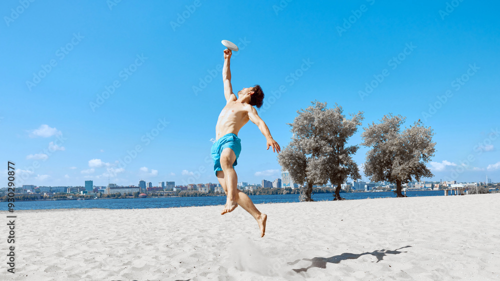 Obraz premium Man in beach shorts playing frisbee on beach by the river under clear blue sky. Dynamic game, catching flying disk in a jump. Concept of sport, summer activity, leisure games, friendship