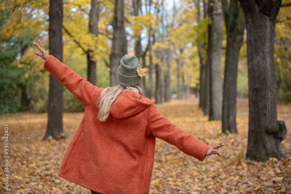 Person in an orange coat and gray beanie with a leaf tucked in their hat walks through a forest with arms outstretched, surrounded by golden autumn leaves, enjoying the peaceful atmosphere.. Person in an orange coat and gray beanie with a leaf tucked in their hat walks through a forest with arms outstretched, surrounded by golden autumn leaves, enjoying the peaceful atmosphere..
