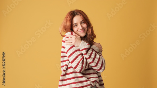 Cute red-haired woman dressed in sweater hugs herself looking at camera on yellow background. Beautiful girl poses in studio with blank space on yellow wall for your promotion photo