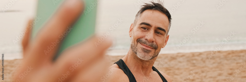Obraz premium Close up, middle-aged athletic man stands on the beach after workout and takes selfie on mobile phone