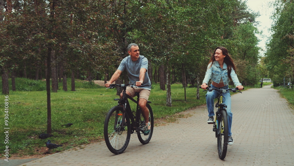 Obraz premium Couple enjoys biking together in park, smiling and talking while riding on tree-lined path. Concept of outdoor activities, bonding, and healthy lifestyle