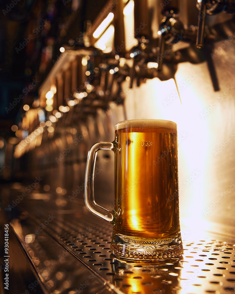 Close-up of frosty mug of golden beer with bubbles, sitting on metallic counter in front of row of beer taps. Concept of beer, brewery, pub, Oktoberfest, traditions Close-up of frosty mug of golden beer with bubbles, sitting on metallic counter in front of row of beer taps. Concept of beer, brewery, pub, Oktoberfest, traditions