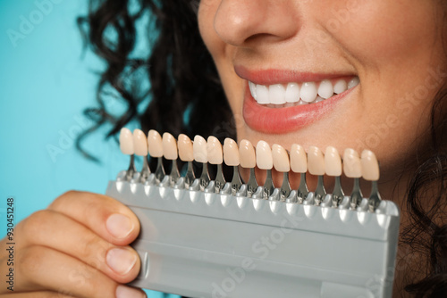 Smiling woman with teeth color samples on light blue background, closeup. Dental veneers