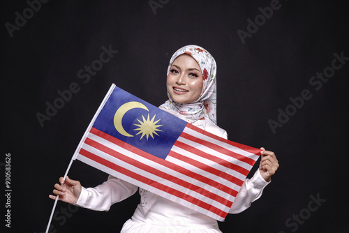 Beautiful Asian girl wearing white gress with hijab, happily holding Malaysian flag isolated over black background. Independence day, new year nationalism concept. photo