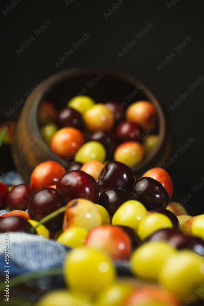 Fototapeta premium Cherries of different colours poured on the table from a clay pot, dark background, close-up