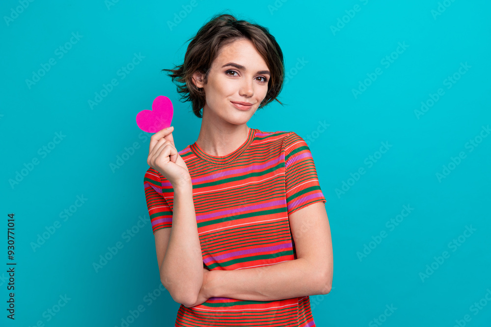 Portrait of satisfied lovely girl with short hair wear stylish t-shirt hold small paper heart in hand isolated on teal color background Portrait of satisfied lovely girl with short hair wear stylish t-shirt hold small paper heart in hand isolated on teal color background