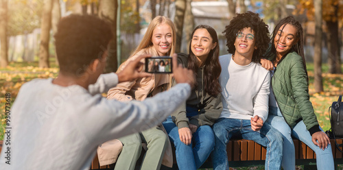Black guy taking photo of his international friends in park, using smartphone, panorama