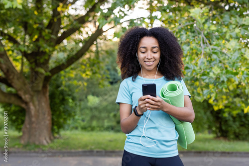 Slika na platnu Smiling woman in park with yoga mat and phone, wearing earphones, enjoying fresh air and fitness motivation