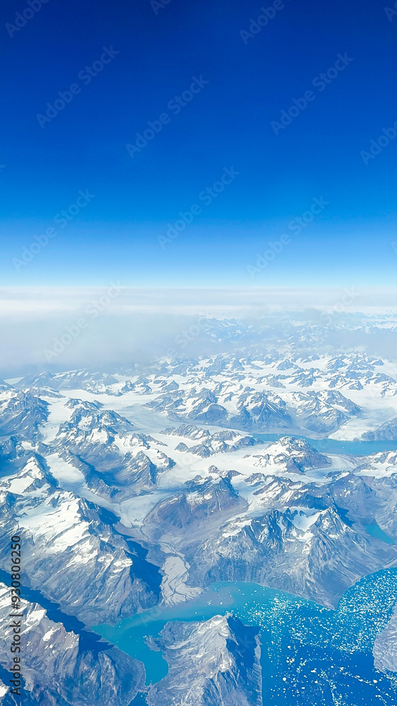 Obraz premium Out of this World Views Flying Over Greenland. Snow-capped mountains, glaciers and pristine terrain. Clear blue skies above the horizon.
