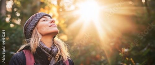 Woman in winter clothing stands in the forest with her eyes closed, breathing deeply and enjoying sunlight filtering through trees. Peaceful and happy expression with a warm golden glow.