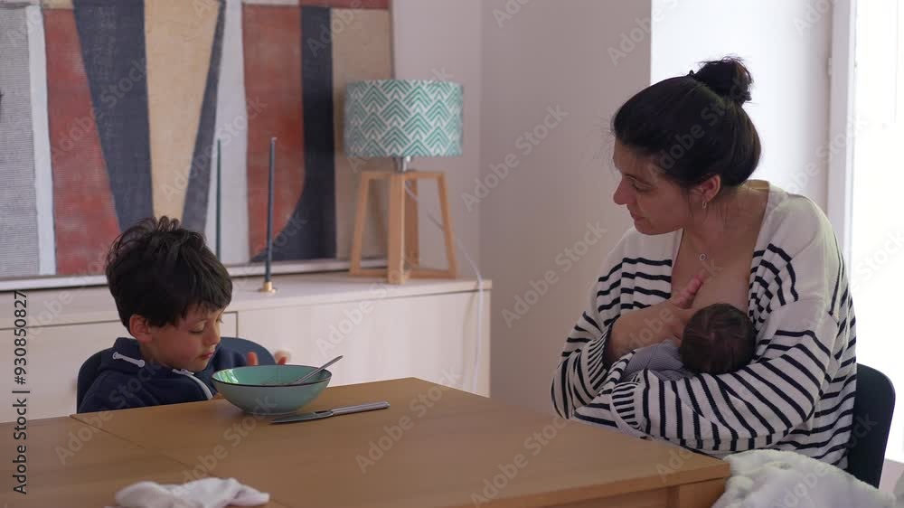 Mother breastfeeding newborn while her older son sits at the dining table, hands on his bowl, seemingly lost in thought, showcasing a quiet, reflective family moment Mother breastfeeding newborn while her older son sits at the dining table, hands on his bowl, seemingly lost in thought, showcasing a quiet, reflective family moment