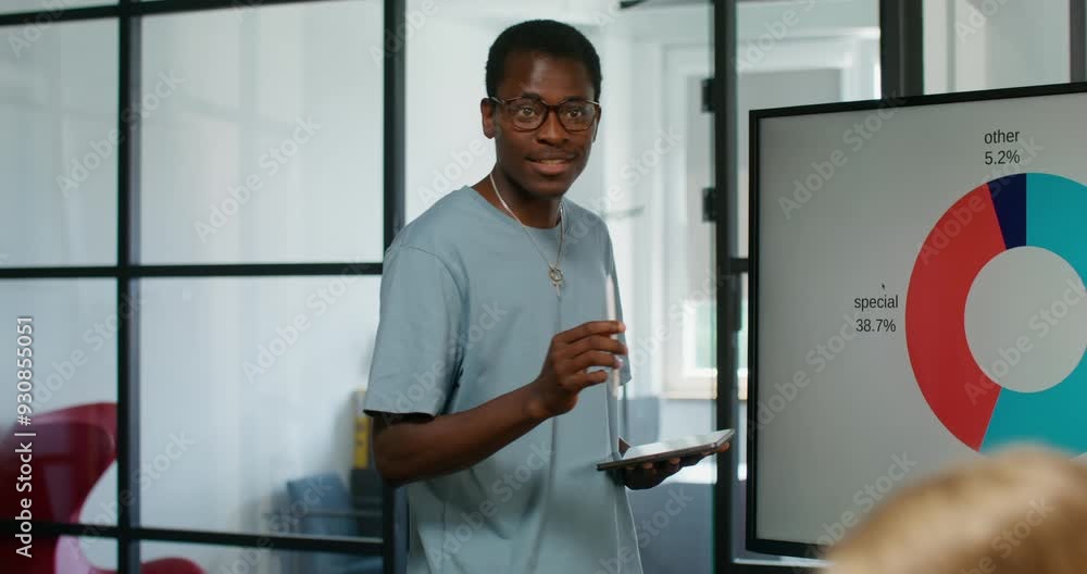 A speaker with a tablet in his hand talks standing in front of his colleagues at an office meeting. Analytical graphs on the screen. Business planning. A speaker with a tablet in his hand talks standing in front of his colleagues at an office meeting. Analytical graphs on the screen. Business planning.