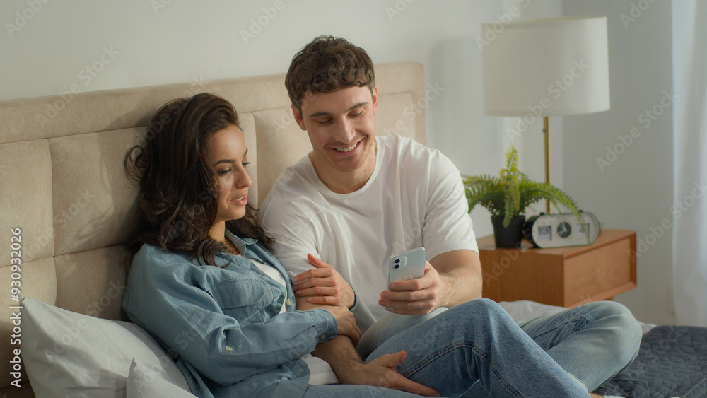 Fototapeta premium Couple man woman bed home bedroom mobile phone smartphone cellphone technology conversation discussion dialogue talk speak together female male browsing online network girlfriend boyfriend relaxing