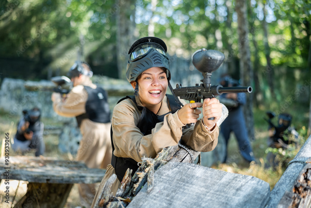 Fototapeta premium Excited caucasian young woman paintball player standing at cover, aiming with paintball gun and shooting.