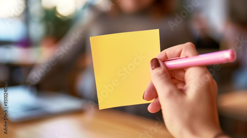 Blank Sticky Note: A close-up shot of a hand holding a bright yellow sticky note with a pink pen, ready to capture thoughts and ideas.  The background is blurred, creating a sense of focus on the note photo