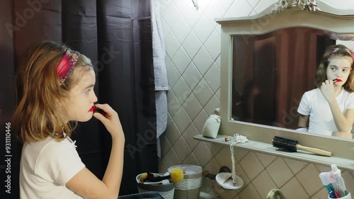 Young girl applies red lipstick in bathroom mirror, smearing it across her cheeks. Her expression shows curiosity and excitement as she explores makeup, creating an endearing and humorous scene. photo