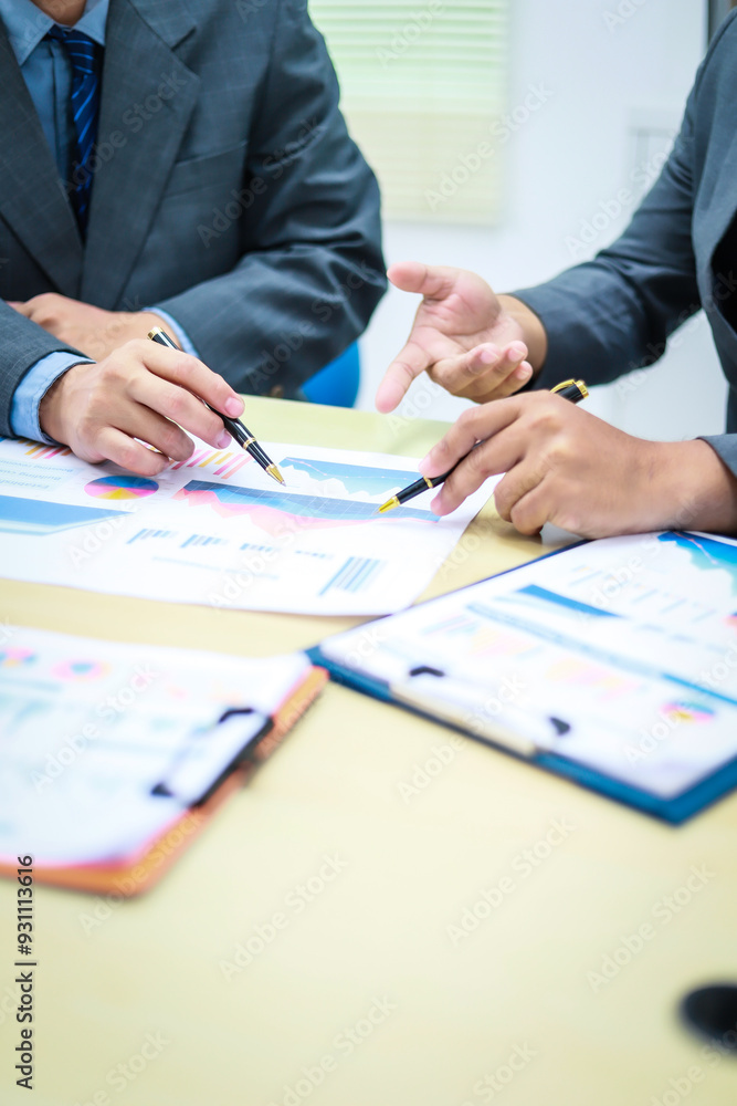 Two businessmen are seated at a desk in an office, engaged in a meeting focused on analysis, finance, and marketing strategies, with documents and graphs spread across the table. Two businessmen are seated at a desk in an office, engaged in a meeting focused on analysis, finance, and marketing strategies, with documents and graphs spread across the table.