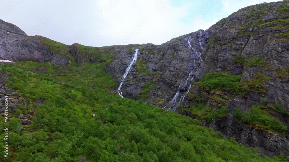 Ominous approaching shot of a waterfall gushing down the rock face of a mountain range in Vassdalsvattnet near Reine in Lofoten Norway. Sky is cloudy, ground is covered in green lush vegetation. Ominous approaching shot of a waterfall gushing down the rock face of a mountain range in Vassdalsvattnet near Reine in Lofoten Norway. Sky is cloudy, ground is covered in green lush vegetation.