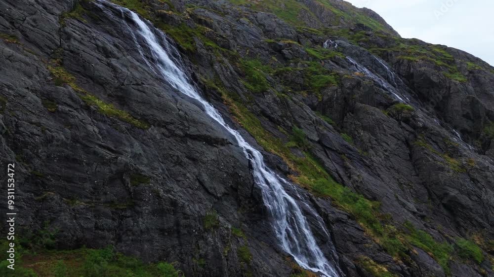 Approaching a long narrow waterfall flowing down rocky cliff. The white water, grey rocks. The scene is peaceful, serene, ominous and majestic. Lofoten mountain range near Reine in northern Norway. Approaching a long narrow waterfall flowing down rocky cliff. The white water, grey rocks. The scene is peaceful, serene, ominous and majestic. Lofoten mountain range near Reine in northern Norway.