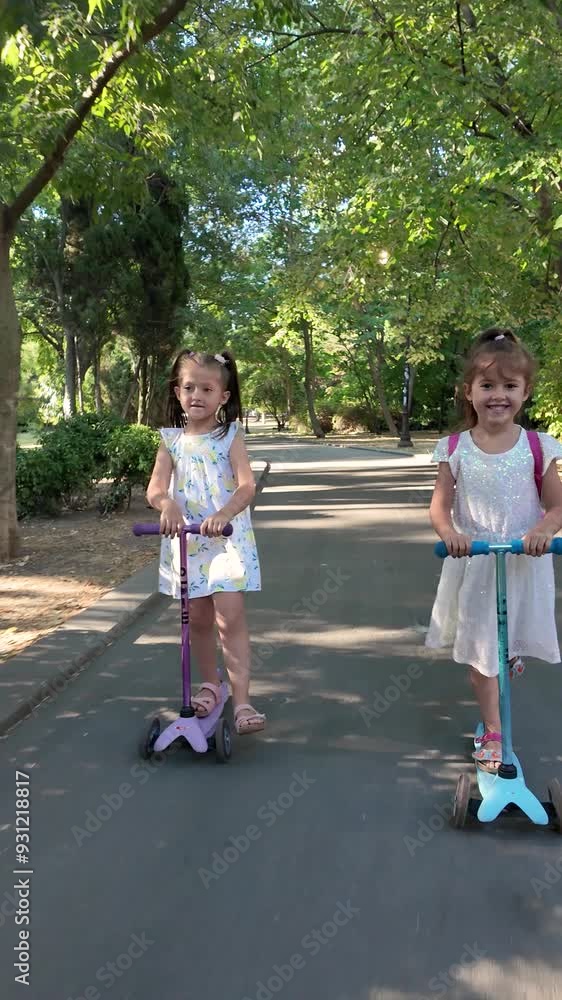 Two girls riding scooters in the park. Kids having fun outdoors with scooters. Vertical video Two girls riding scooters in the park. Kids having fun outdoors with scooters. Vertical video