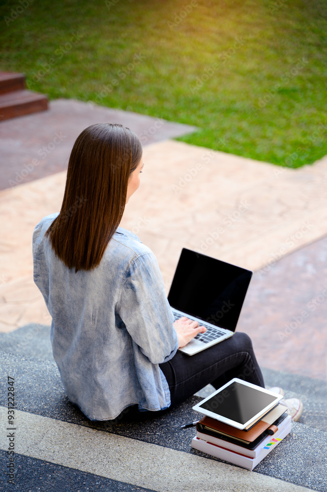 Obraz premium A young woman working on a laptop while sitting outdoors on a stone staircase surrounded by books and a tablet in university