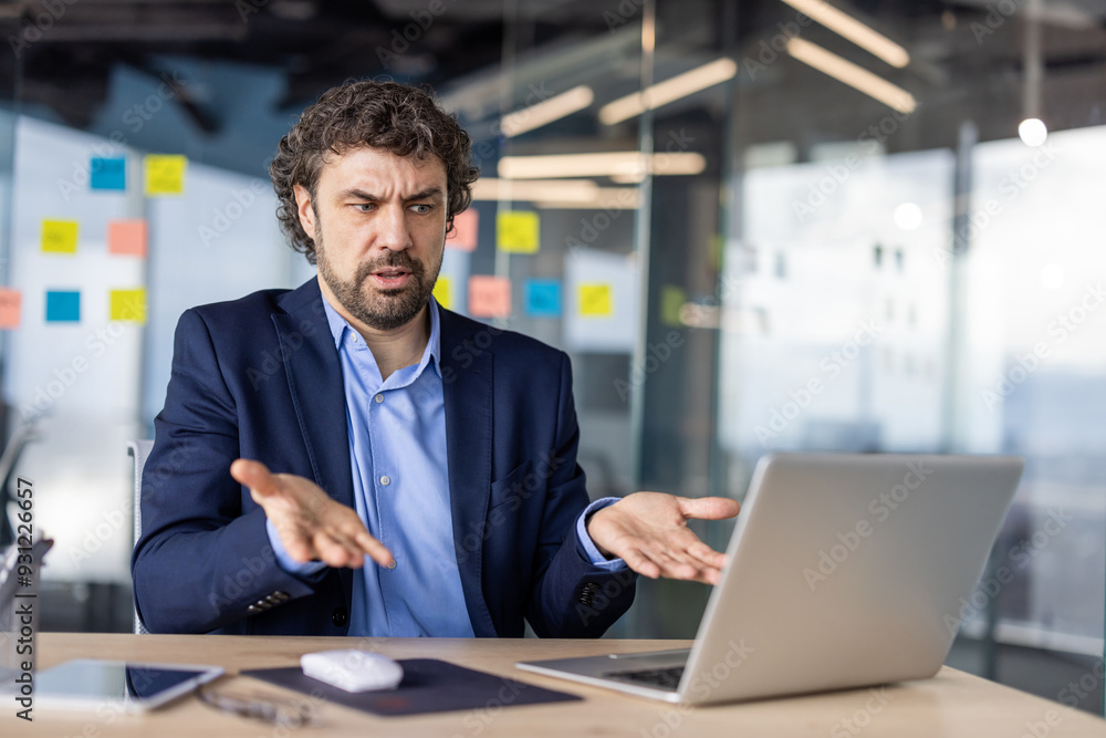 A businessman in a suit shows confusion and frustration at his laptop screen in an office with sticky notes on the walls. A businessman in a suit shows confusion and frustration at his laptop screen in an office with sticky notes on the walls.