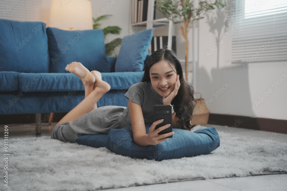 Obraz premium Young woman is lying on her stomach on the floor, using her smartphone and smiling
