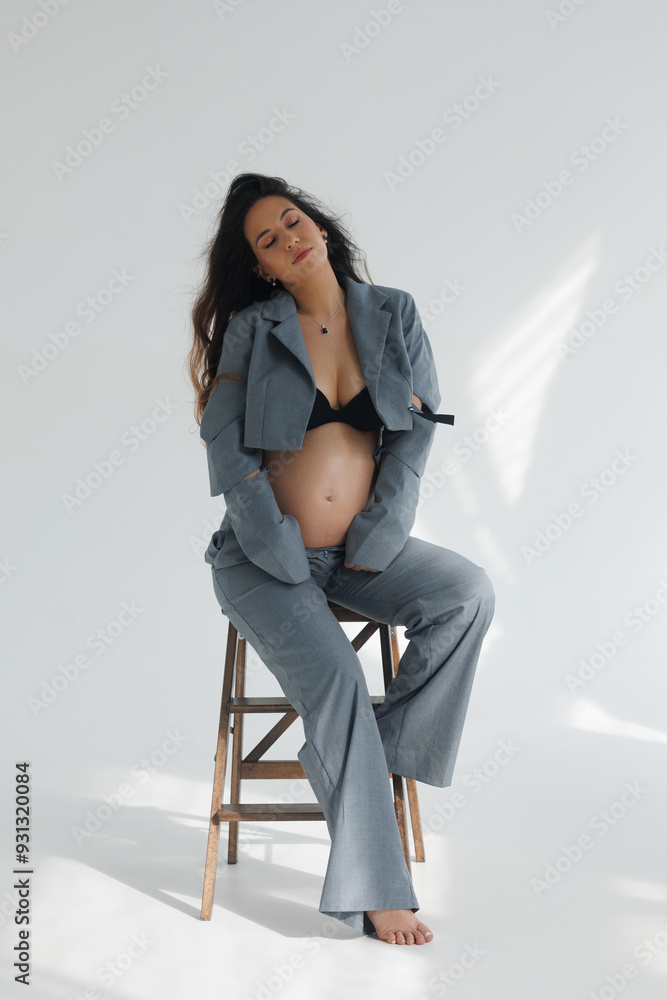 A beautiful pregnant brunette woman in a cropped gray stylish suit on a white background in the studio. A pretty girl with a bare stomach in a fashionable suit is sitting on wooden staircase barefoot. A beautiful pregnant brunette woman in a cropped gray stylish suit on a white background in the studio. A pretty girl with a bare stomach in a fashionable suit is sitting on wooden staircase barefoot.