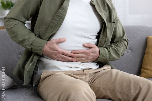 Man sitting on couch experiencing abdominal pain holding stomach. Wearing casual clothing and showing visible discomfort. Concept of stomachache, digestive issues, or health concerns.