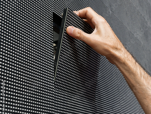 A man's hand removing a square LED module from its place on the surface of a large LED screen for repair and maintenance