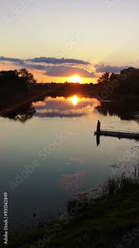 Sunset over the Khorol River in Myrhorod, Poltava region, Ukraine. Human silhouette on a pier, as orange and purple clouds and setting sun reflect on the calm water. Vertical 4K video 29 seconds photo