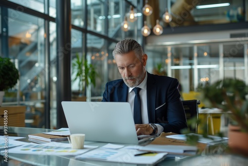 A successful businessman in formal wear, sitting at his desk, working on a laptop in a modern office.