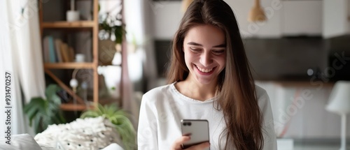 Wallpaper Mural A smiling young woman with long hair enjoys using her smartphone in bright, cozy home. The modern, airy interior features warm lighting, plants, and shelves, creating a relaxed and inviting atmosphere Torontodigital.ca
