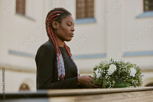 Young African American woman dressed in total black holding rose bouquet before putting it down on coffin