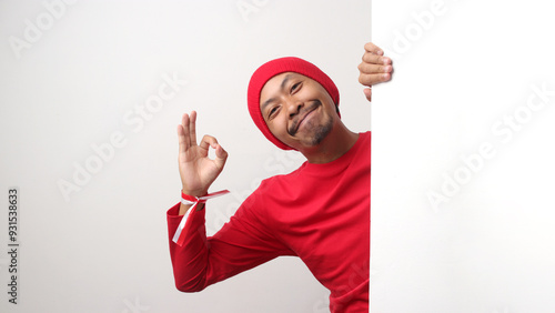 Indonesian man in a red Long Sleeve T-shirt and beanie hat shows the OK gesture to the camera behind a white wall during Indonesia Independence Day. The scene is isolated on a red background. photo