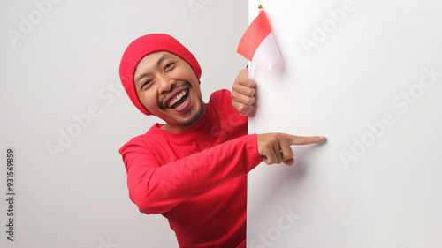 Indonesian man in a red T-shirt and beanie points at copy space, promoting a special offer or discount during Indonesia Independence Day. He stands behind a white wall, isolated on a white background photo