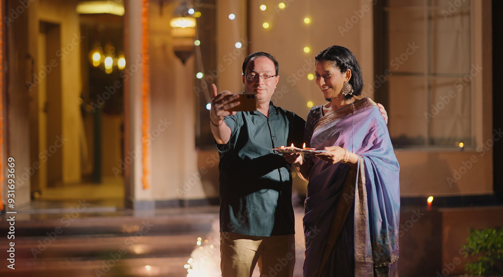 Obraz premium Indian Asian Hindu happy elder old gen x couple hold phone use cam look cute take pose selfie shoot video decor light home older aged male man female wife lady mom dad enjoy fun joy Diwali night time