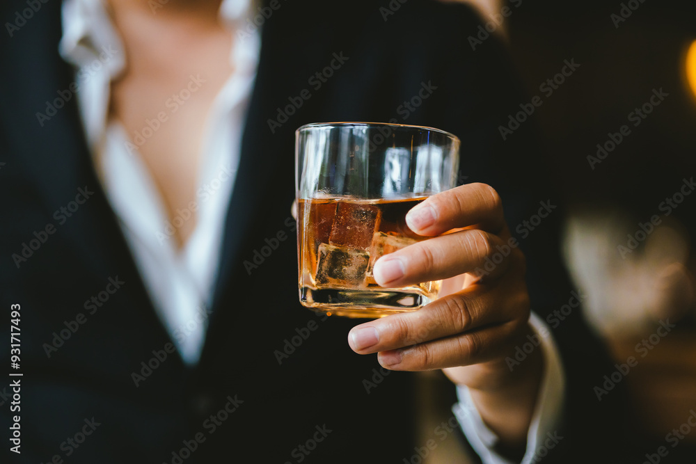 A stylish man holds a glass of whiskey in a fancy pub amidst a crowd of celebrants. The warm and elegant atmosphere accentuates the rich amber-colored liquid. A stylish man holds a glass of whiskey in a fancy pub amidst a crowd of celebrants. The warm and elegant atmosphere accentuates the rich amber-colored liquid.