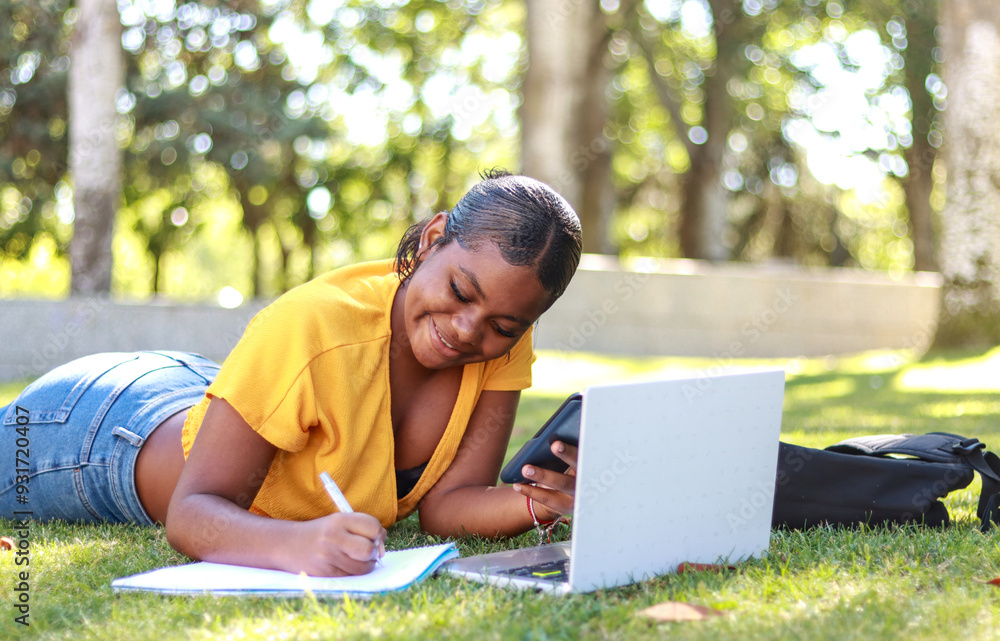 Hispanic woman student lying on grass with a laptop, smartphone, and notebook, studying outdoors and smiling as she completes her schoolwork Hispanic woman student lying on grass with a laptop, smartphone, and notebook, studying outdoors and smiling as she completes her schoolwork