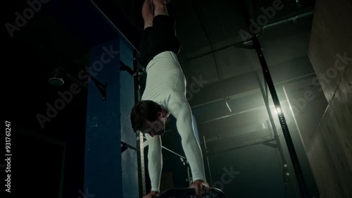 Professional male gymnast using bars while performing handstand with legs apart at dark loft style sports studio photo